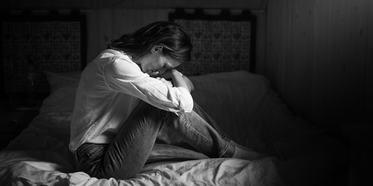 Black and white photo of an upset woman in her room. Sitting on her bed.